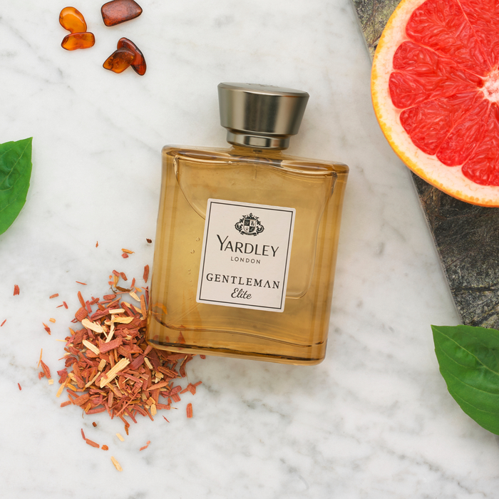Yardley London Gentleman Elite amber glass bottle on white marble, surrounded by wood shavings, grapefruit slice, leaves and amber resin pieces.
