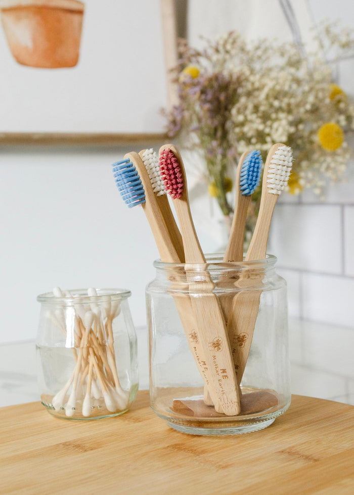 Set of bamboo toothbrushes in a glass jar on a wooden surface with a white tiled wall in the background.