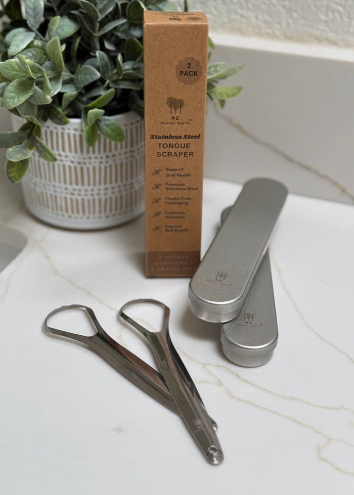 Two stainless steel tongue scrapers on a marble surface with a cardboard box labeled '2 Pack' in the background.