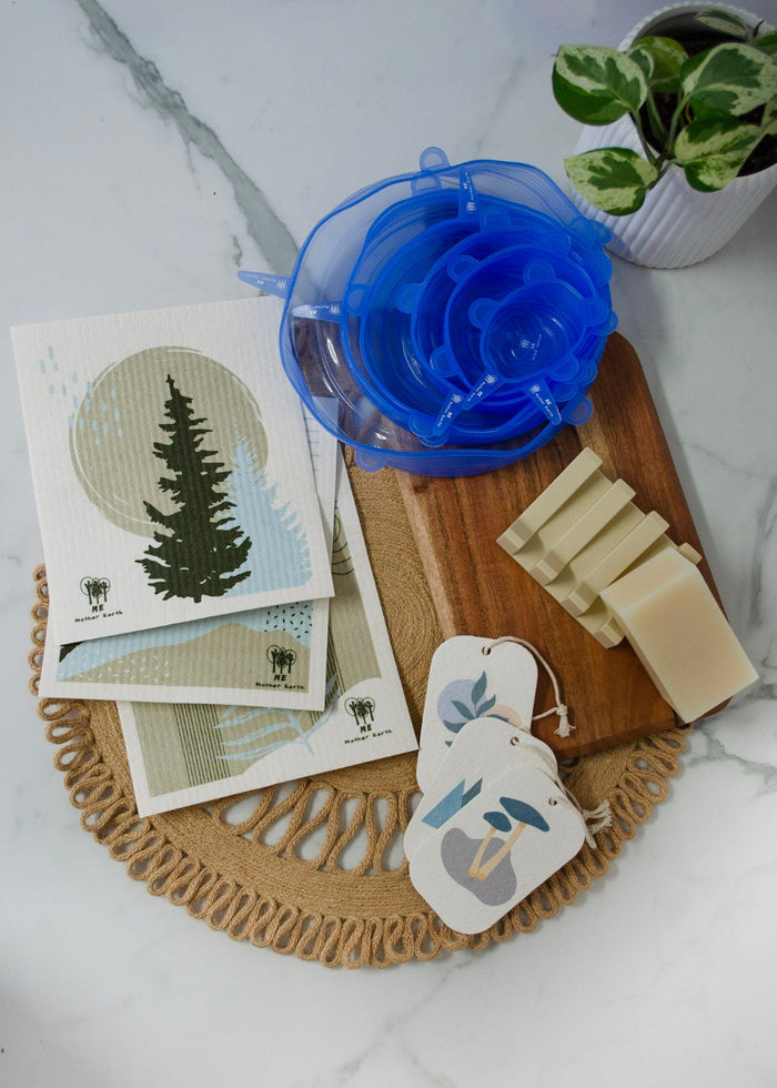Wooden cutting board with soap, cellulose sponges, and a blue silicone bowl covers on a marble surface.