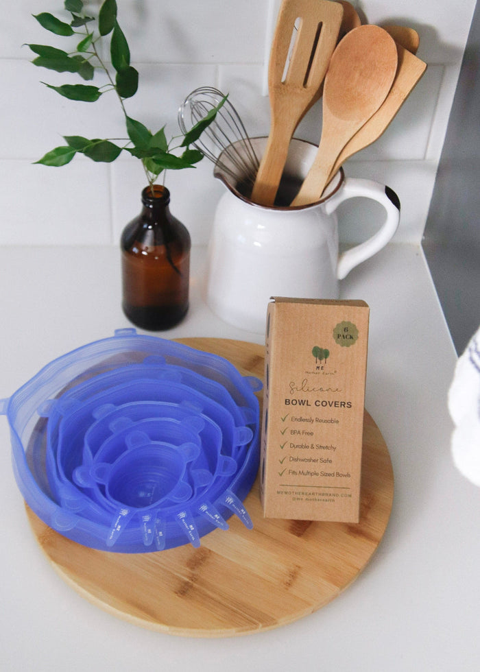 Blue silicone bowl cover on a wooden board with kitchen utensils and a bottle in the background.