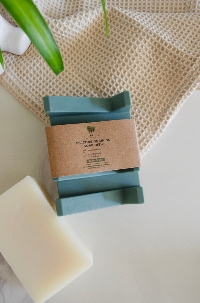 A silicone draining soap dish in green, placed on a white surface with a burlap background, next to a bar of soap and a plant leaf.