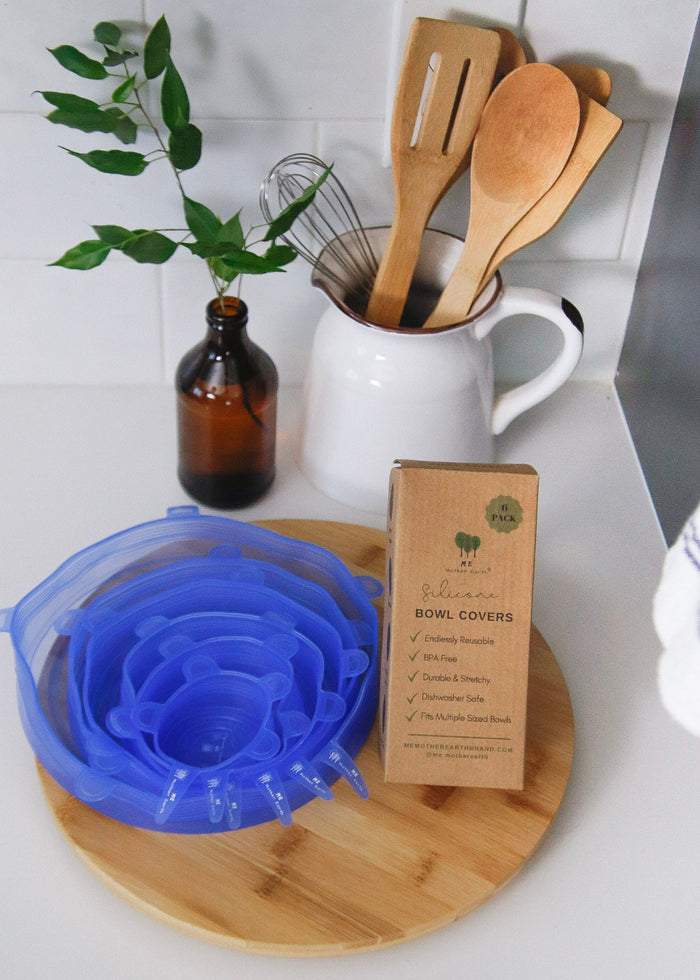 Kitchen scene with a blue silicone bowl cover, wooden utensils, and a plant on a white countertop.