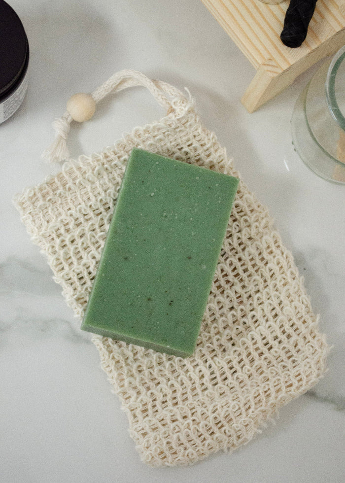 Green bar of soap on a natural fiber soap bag on a light surface.