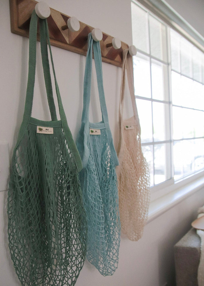 Three mesh shopping bags in green, blue, and beige hanging on a wooden rack by a window.