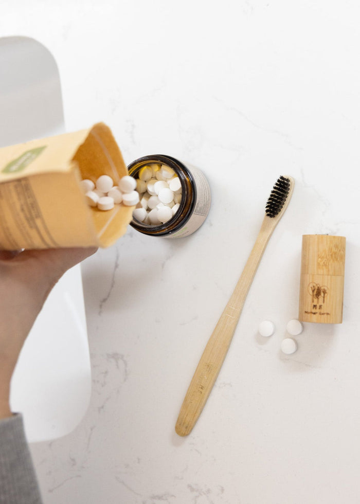 Person holding a box of dental tablets next to a bamboo toothbrush and small container on a white surface.