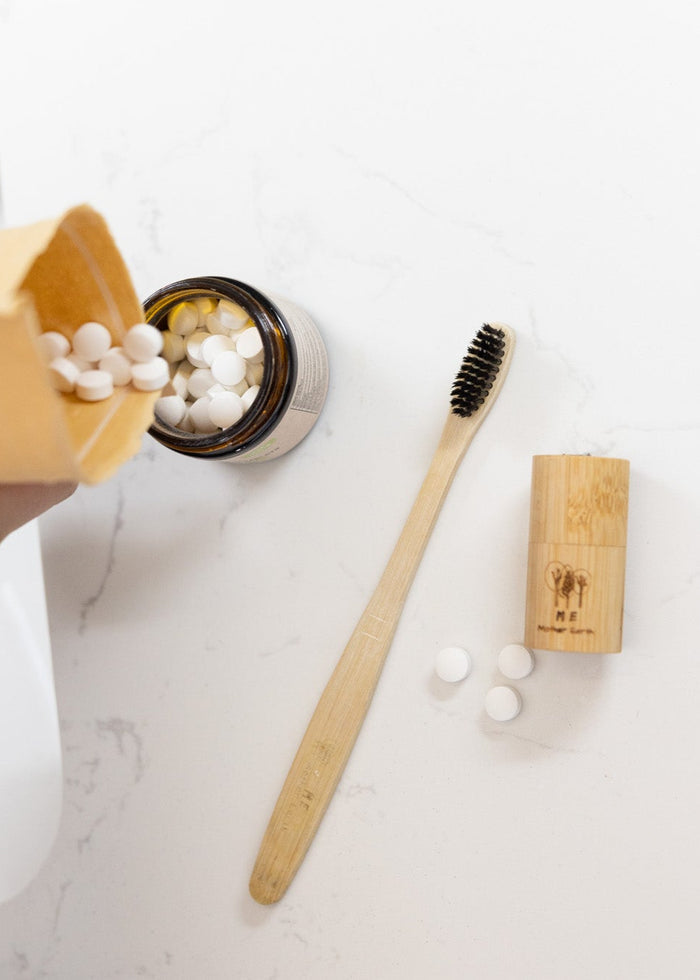 Person holding a container of beads over a small container on a white surface with a bamboo toothbrush and holder.